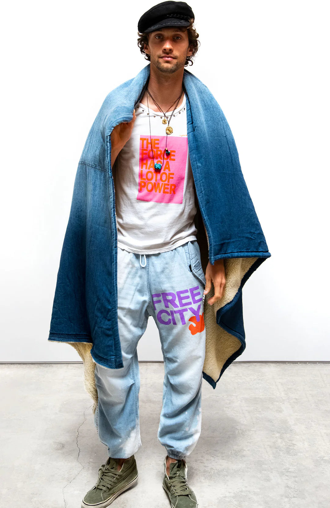 A man stands against a plain background, wearing a dark cap, white shirt with “THE FORCE HAS A LOT OF POWER” in orange and pink, light blue FREE CITY sweatpants, green sneakers, and the Free City Indigo Sherpa Blanket/Indigo Blues.