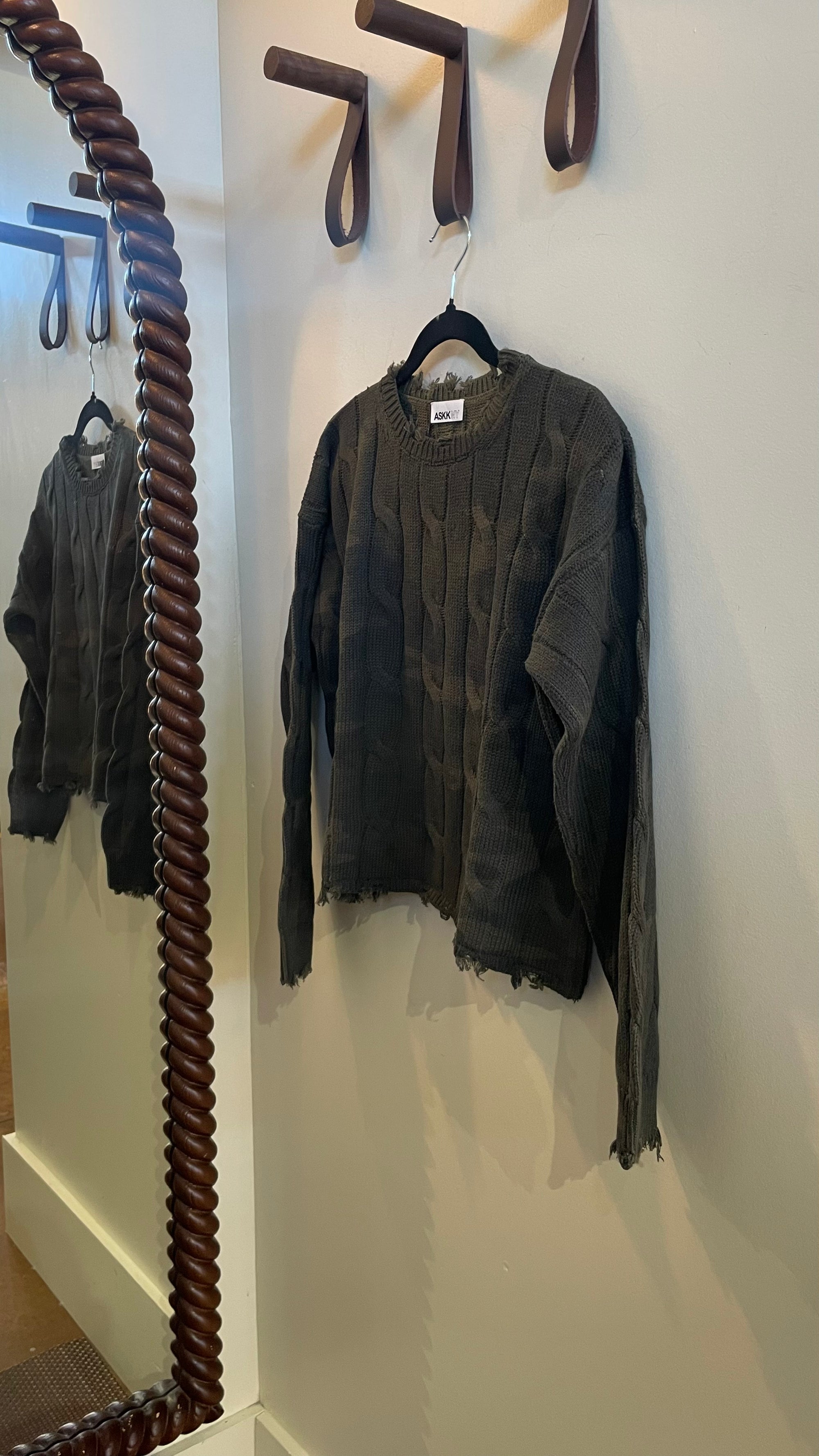ASKK Cable Crop Crew dark olive green cable-knit camo sweater with frayed hem hanging on a wall hook.