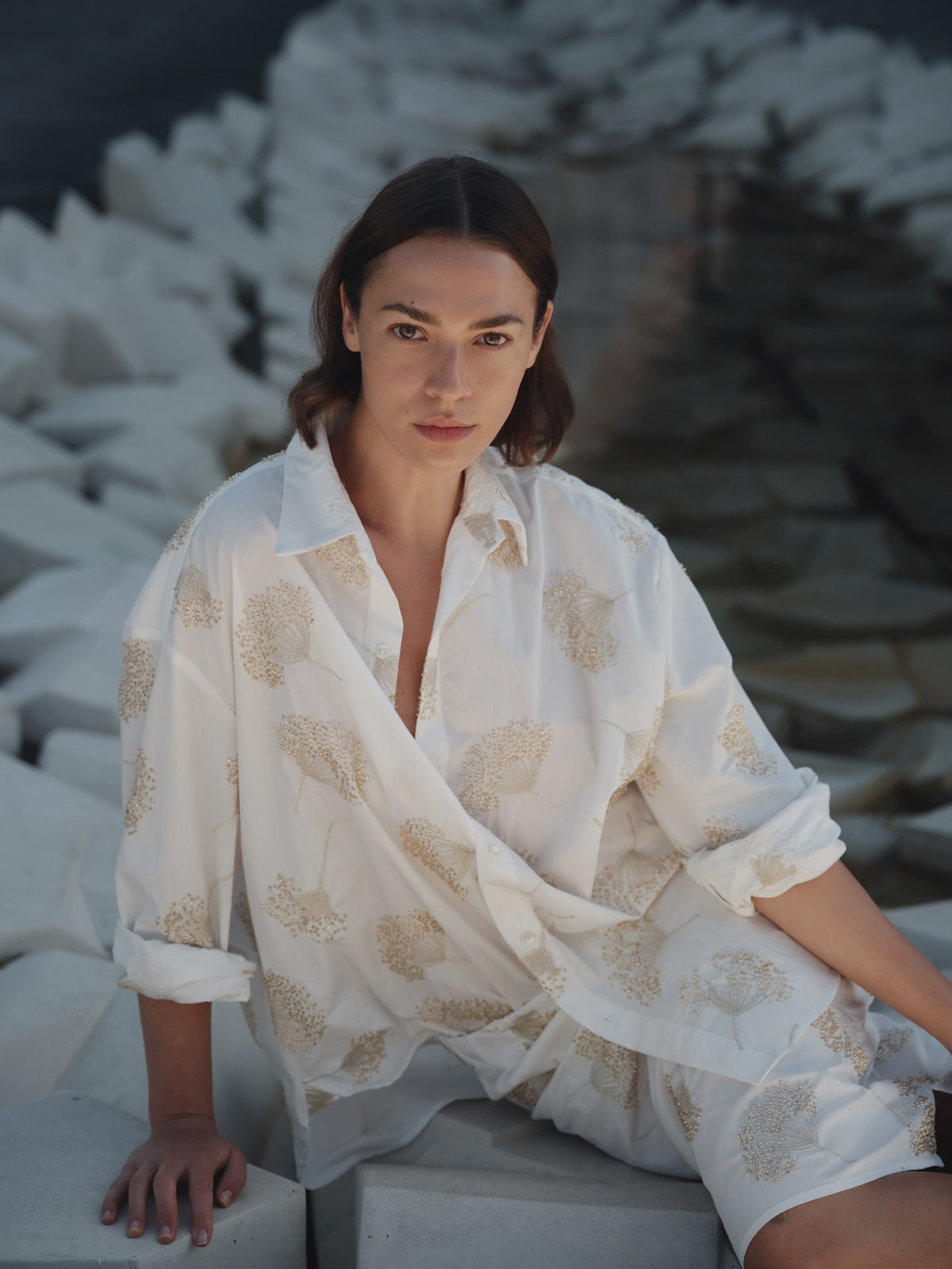A woman with shoulder-length brown hair sits on angular white stones, wearing Cissa's The Mr Big Shirt—a loose cotton shirt with a beige floral pattern and adjustable cuffs—looking calmly at the camera.