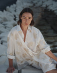 A woman with shoulder-length brown hair sits on angular white stones, wearing Cissa's The Mr Big Shirt—a loose cotton shirt with a beige floral pattern and adjustable cuffs—looking calmly at the camera.