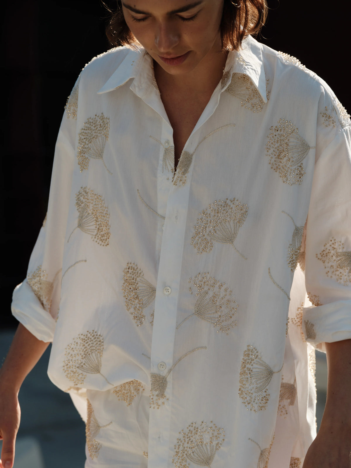 A person wears The Mr Big Shirt by Cissa—a loose cotton shirt with a beige floral pattern and adjustable cuffs. Sleeves are rolled up and only the upper body is visible, set against a softly blurred background.