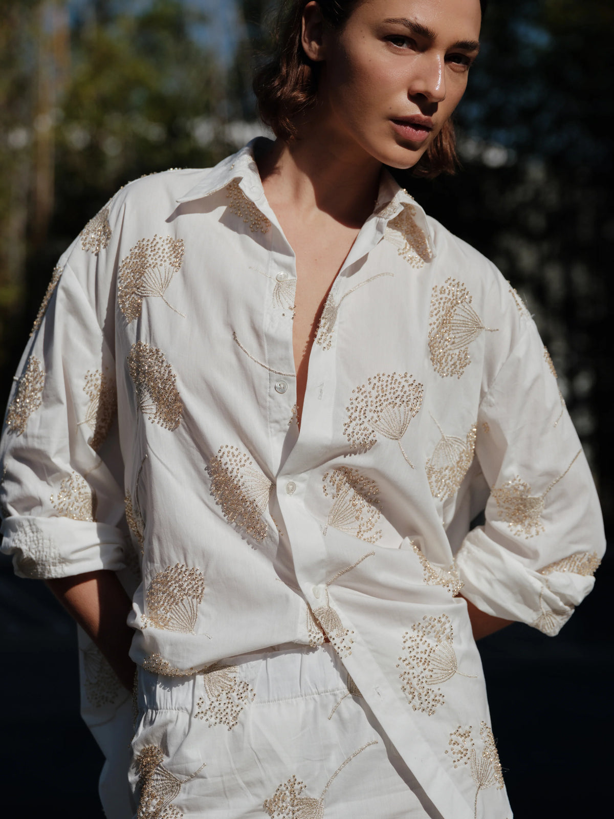 A person stands outdoors in the Cissa Mr Big Shirt—a loose, white cotton shirt with adjustable cuffs—and matching beige floral pants, hands in pockets as sunlight highlights their face and upper body.
