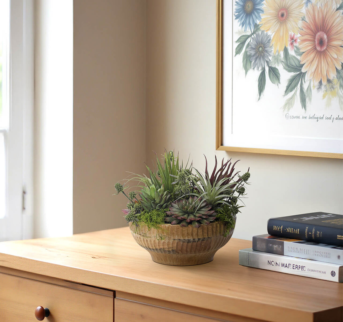 The Succulent Wood Bowl by Natural Decorations Inc. sits on a wooden dresser beside books and a floral painting, bathed in natural light—an ideal, maintenance-free accent for any cozy space.