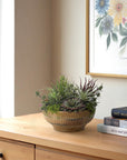 The Succulent Wood Bowl by Natural Decorations Inc. sits on a wooden dresser beside books and a floral painting, bathed in natural light—an ideal, maintenance-free accent for any cozy space.