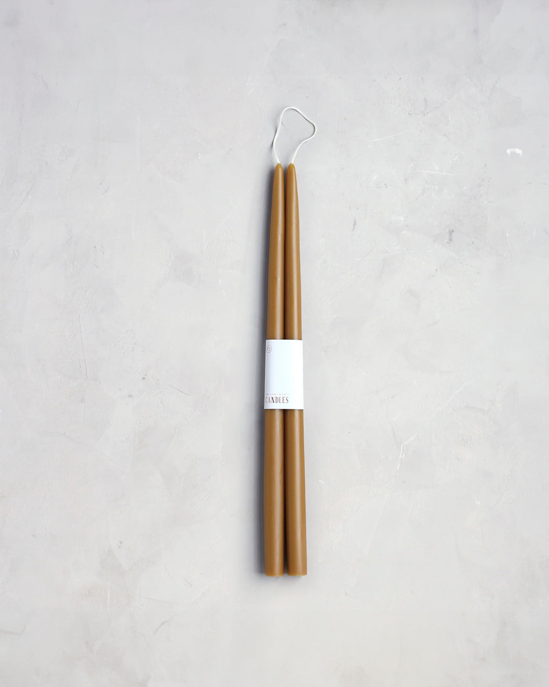 Two tall, 18" taper candles from The Floral Society stand upright against a light gray, textured background. Wrapped near their centers with a white label featuring minimalist text, these 18" Taper Candle - Pair exude a clean and sleek appearance, perfect for adding a graceful ambiance to any setting.