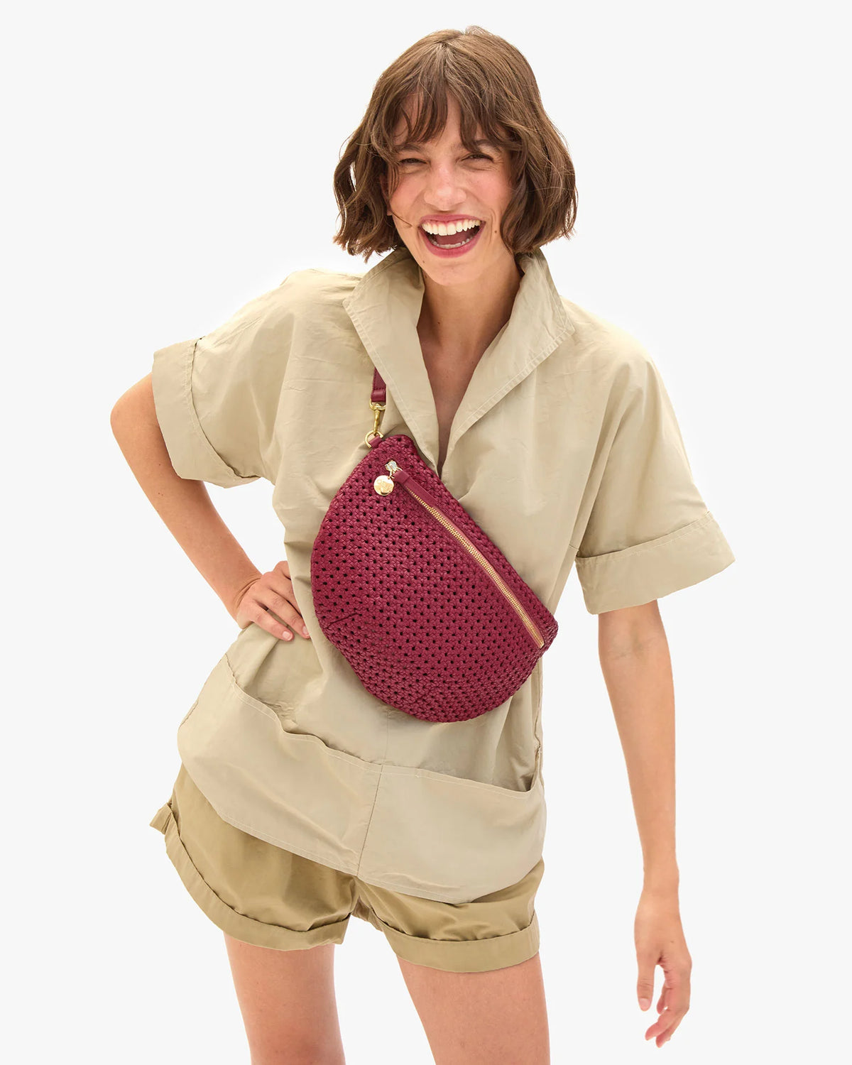 A smiling woman with short brown hair wears a beige short-sleeve shirt and shorts, accessorized with the Clare Vivier Grande Fanny S26 crossbody bag. She stands against a plain white background.