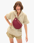 A smiling woman with short brown hair wears a beige short-sleeve shirt and shorts, accessorized with the Clare Vivier Grande Fanny S26 crossbody bag. She stands against a plain white background.