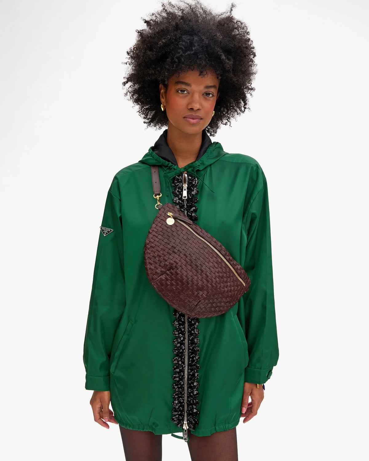 A woman with curly hair, wearing a green hooded jacket with black trim, carries the Clare Vivier Grande Fanny S26—a large handwoven leather bag with an adjustable strap—against a plain white background.