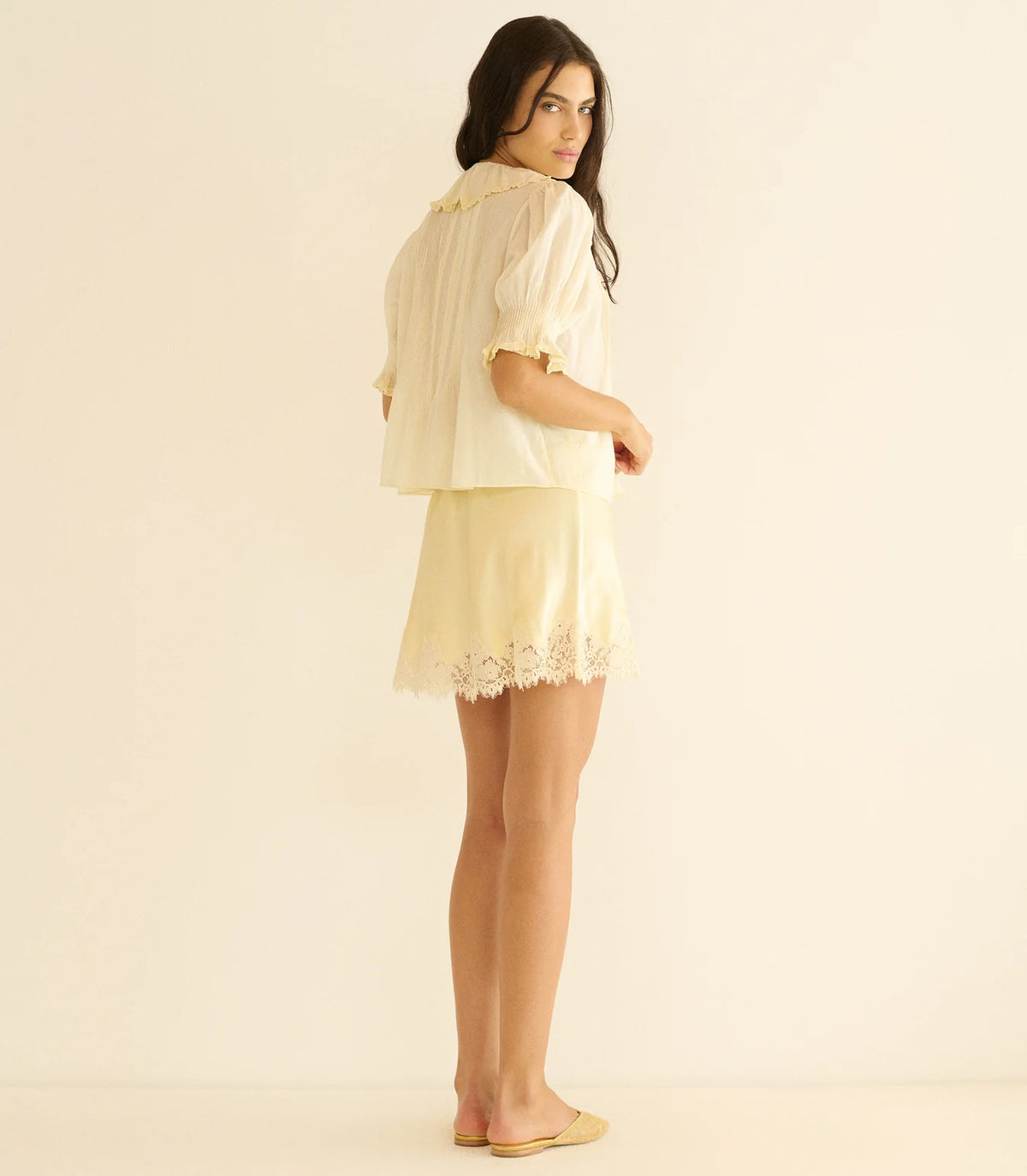 A woman with long dark hair stands sideways, barefoot, against a plain light background, wearing the light yellow DOEN Henri Top with short sleeves and lace trim, paired with a matching skirt.