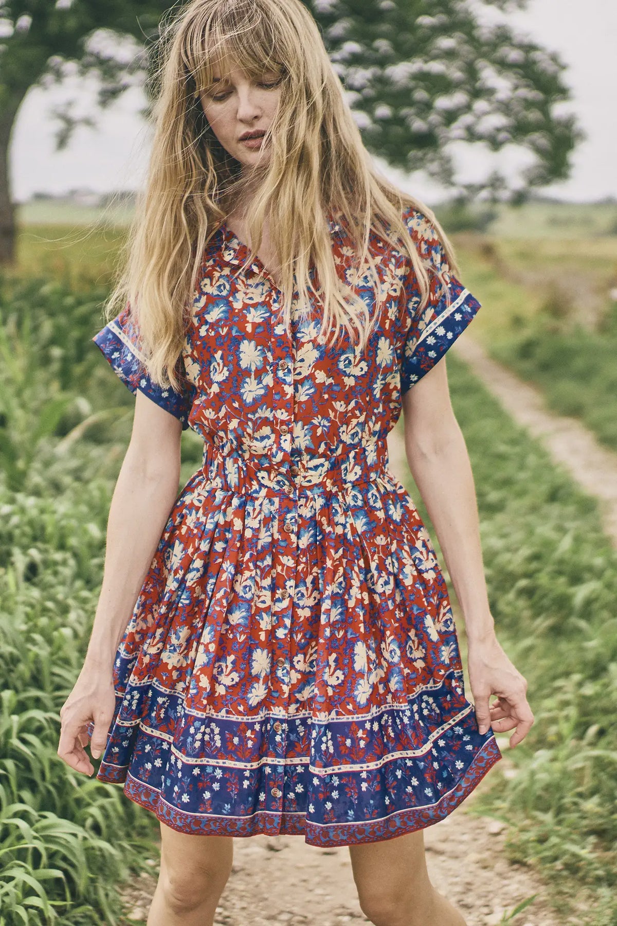 A woman with long blonde hair wears the Howard Dress by Hunter Bell NYC, featuring a shirred skirt, as she stands on a dirt path surrounded by green grass and trees on a cloudy day.