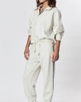 Dressed in Ragabond Studio's off-white Studio Sweats Checkerboard jogger pants with a black checkered stripe, this person completes the look with black loafers while standing against a plain white background.