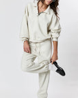 Wearing Ragabond Studio’s Studio Sweats Checkerboard and matching pullover, a person stands on one leg with a tilted head, touching an ankle, against a plain white background.