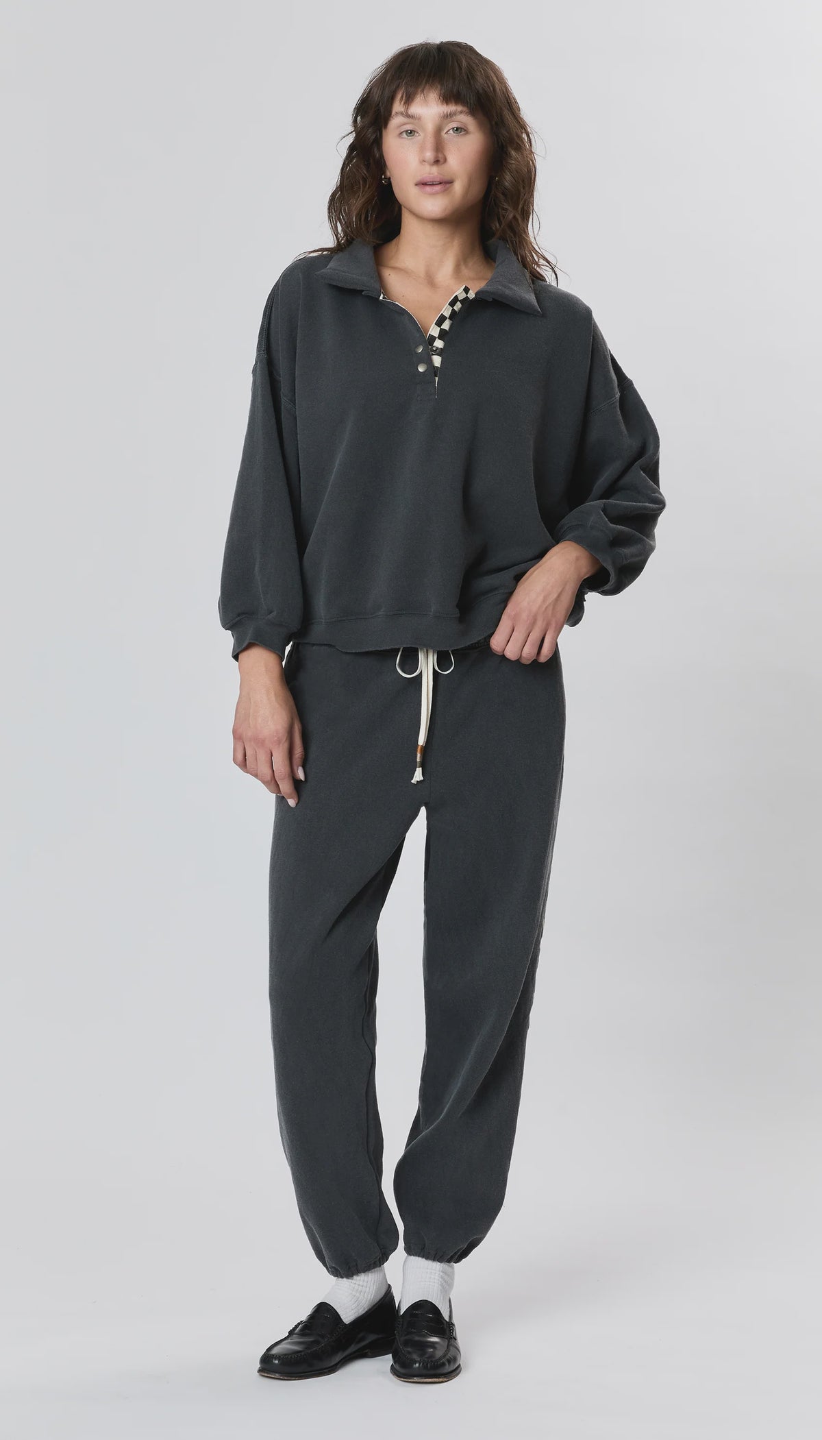 A person stands facing forward in a loose dark gray pullover and Ragabond Studio’s Studio Sweats Checkerboard jogger pants. They have medium-length brown hair, black shoes with white socks, and a plain light background.