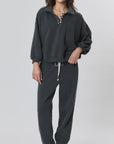 A person stands facing forward in a loose dark gray pullover and Ragabond Studio’s Studio Sweats Checkerboard jogger pants. They have medium-length brown hair, black shoes with white socks, and a plain light background.