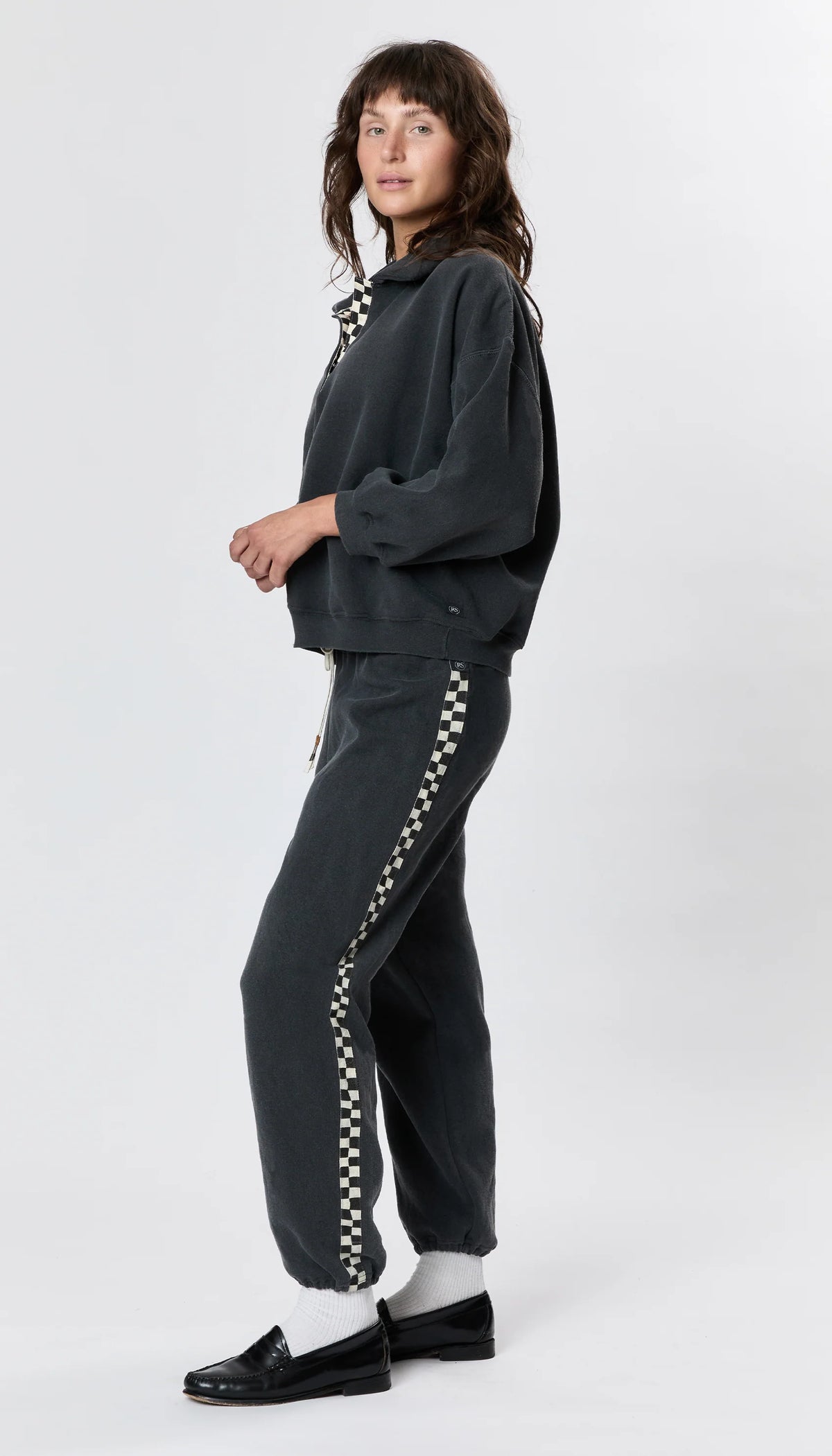 A person stands sideways in Ragabond Studio’s Studio Sweats Checkerboard, featuring a black-and-white checkerboard stripe, paired with black shoes and white socks against a plain light background.