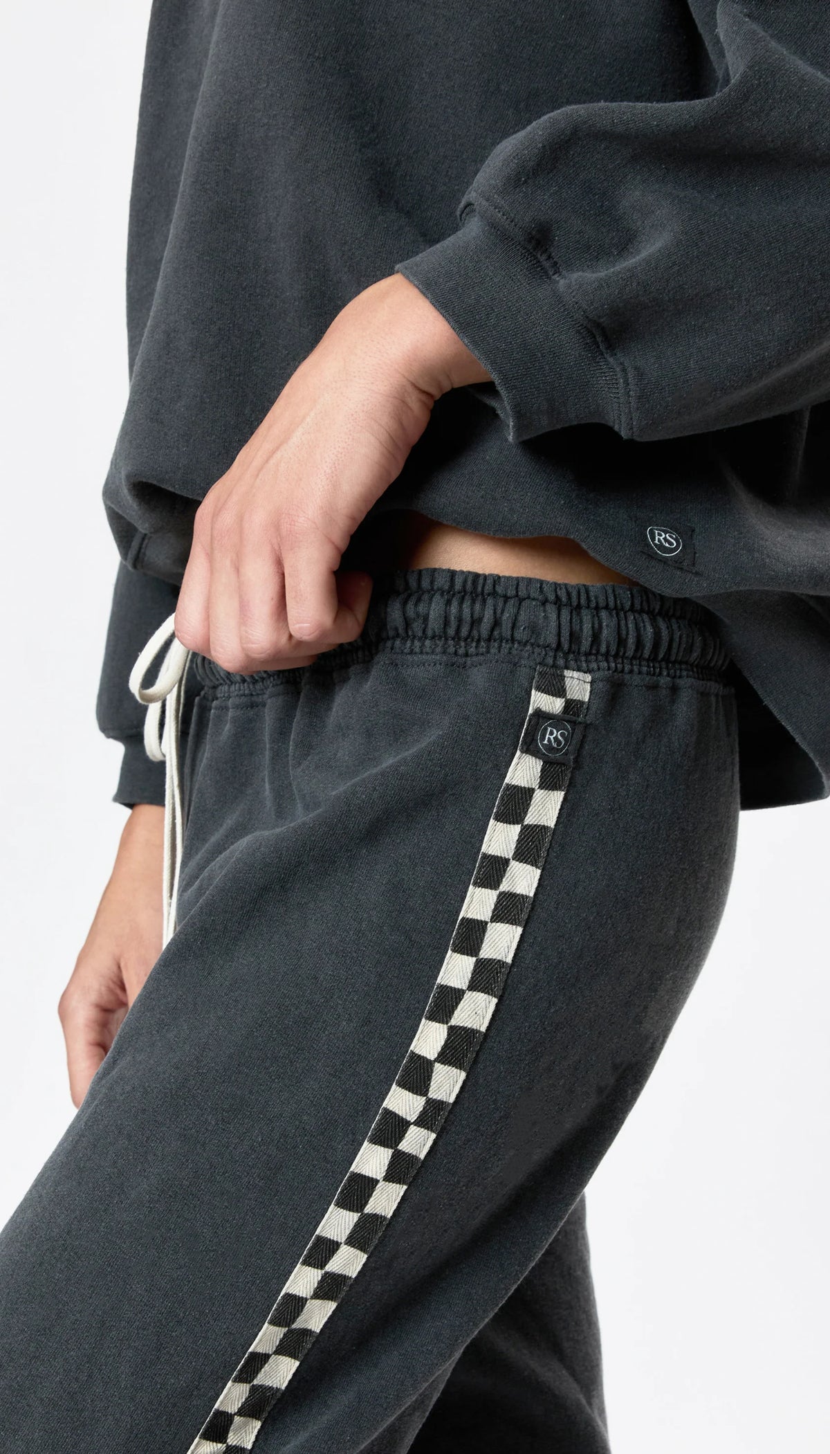 A person wears Ragabond Studio’s Studio Sweats Checkerboard, featuring a black and white checkerboard side stripe, paired with a matching dark sweatshirt. Only their torso and upper legs are visible.