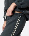 A person wears Ragabond Studio’s Studio Sweats Checkerboard, featuring a black and white checkerboard side stripe, paired with a matching dark sweatshirt. Only their torso and upper legs are visible.