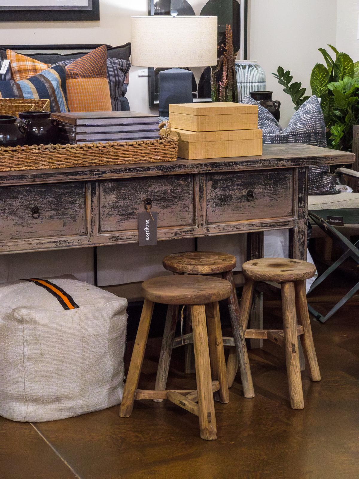 A rustic wooden table with four drawers, adorned with a basket, a lamp, and decorative items sits in the space. Underneath are three imported ROUND STOOLs by Indus Design Imports that add a handmade vintage charm. The area is also decorated with a white pouf and various plants. In the background, pillows and furniture can be seen, enhancing the cozy atmosphere with timeless elegance.