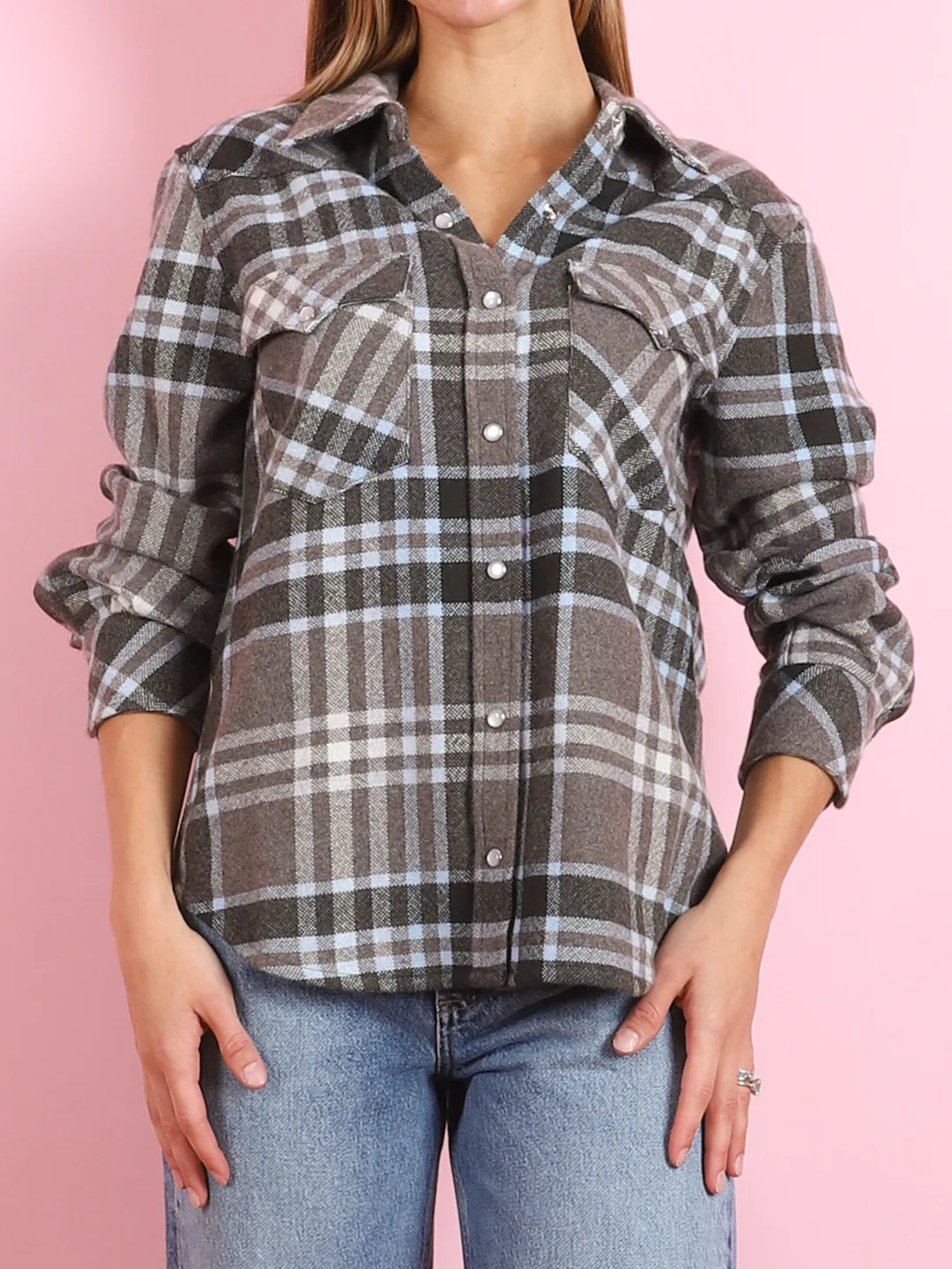 A person wears the KUJTEN Lumber L/S Shirt, a gray plaid button-up with rolled sleeves, light blue jeans, and a cozy women's sweater, standing against a pink background.