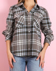 A person wears the KUJTEN Lumber L/S Shirt, a gray plaid button-up with rolled sleeves, light blue jeans, and a cozy women's sweater, standing against a pink background.