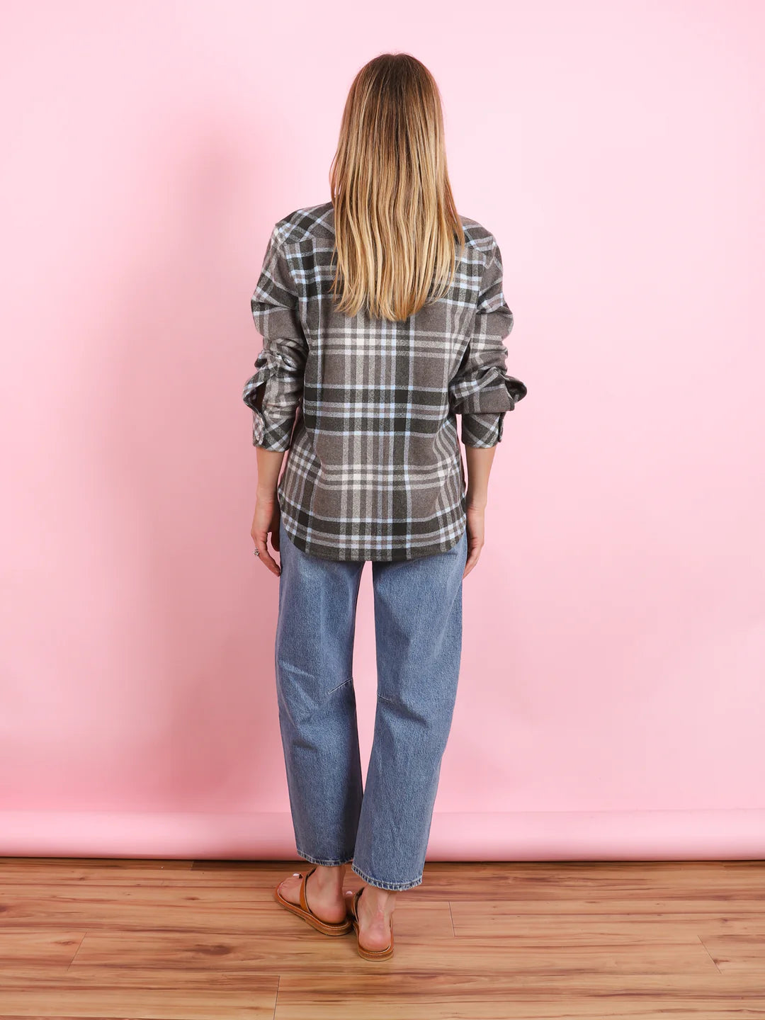 A person with long blonde hair stands facing away, wearing the KUJTEN Lumber L/S Shirt in gray plaid, blue jeans, and sandals, set against a pink background and wooden floor.