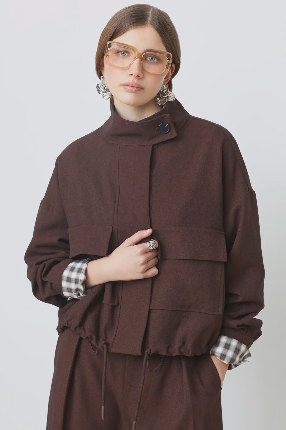 SUNCOO Dona jacket layered over a plaid shirt and brown pants, styled with silver earrings and glasses.