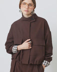 SUNCOO Dona jacket layered over a plaid shirt and brown pants, styled with silver earrings and glasses.