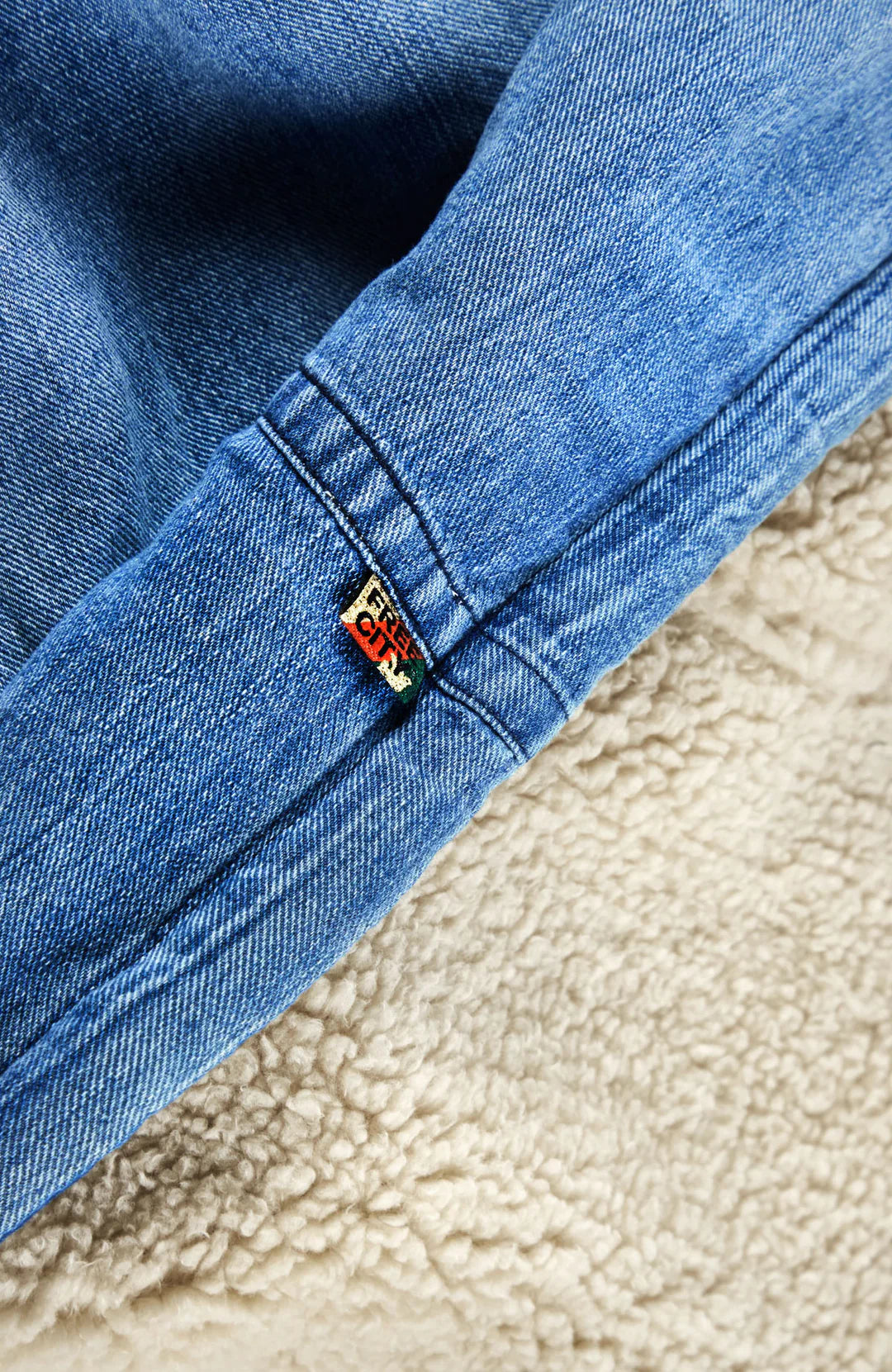 Close-up of the Indigo Sherpa Blanket/Indigo Blues by Free City (sparrow, LLC), showing blue Japanese indigo denim with a multicolored tag near the seam, lying atop soft beige SUPERFLUFF sherpa fleece.