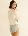A woman with long brown hair wears the DOEN Iona Short—light blue, high-waisted with delicate floral lace trim—and a soft cream sweater, standing sideways and looking over her shoulder against a plain light background.