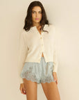A woman with long brown hair wears the DOEN Iona Short and a cream buttoned cardigan, standing against a plain beige background.