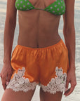 A person stands on a sandy beach, wearing a green floral bikini top paired with the DOEN Iona Short, featuring high-waist silk satin and white lace trim. The ocean appears blurred in the background.