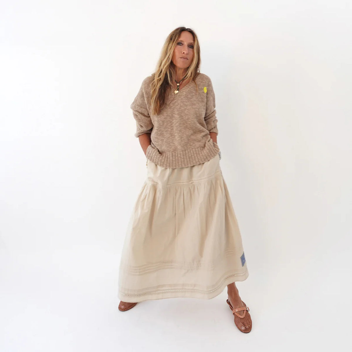 A woman with long hair stands against a white background wearing the Kerri Rosenthal Summerland V Neck Sweater, paired with a long cream skirt and brown sandals. Her hands are in her pockets.