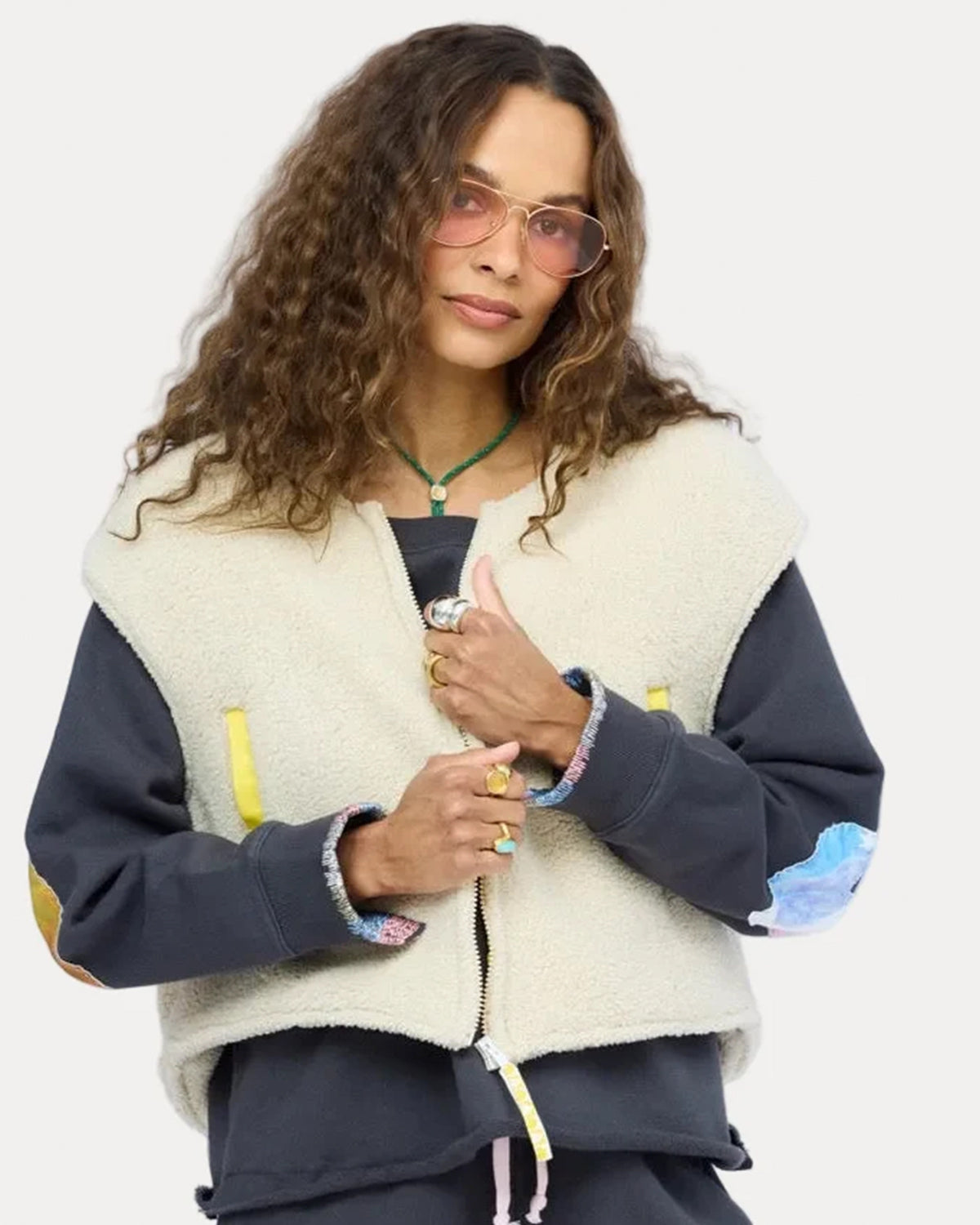 Kerri Rosenthal Coze Magoze Vest styled with a dark patched sweatshirt and pink glasses, worn by a woman with curly hair.