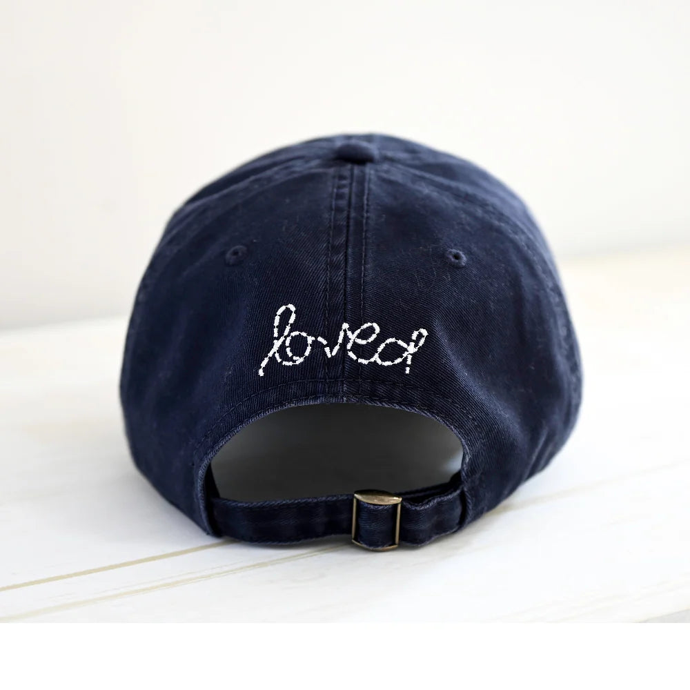The Kerri Rosenthal Baseball Hat Heart Patch features "loved" and a white embroidered heart above the adjustable strap—a navy cap and chic essential, shown here on a light surface.