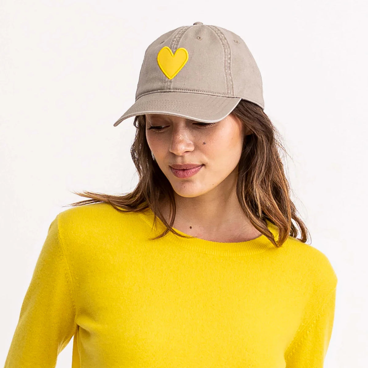 A woman smiles softly, looking down while wearing a Kerri Rosenthal Baseball Hat Heart Patch and a bright yellow sweater against a plain white background.