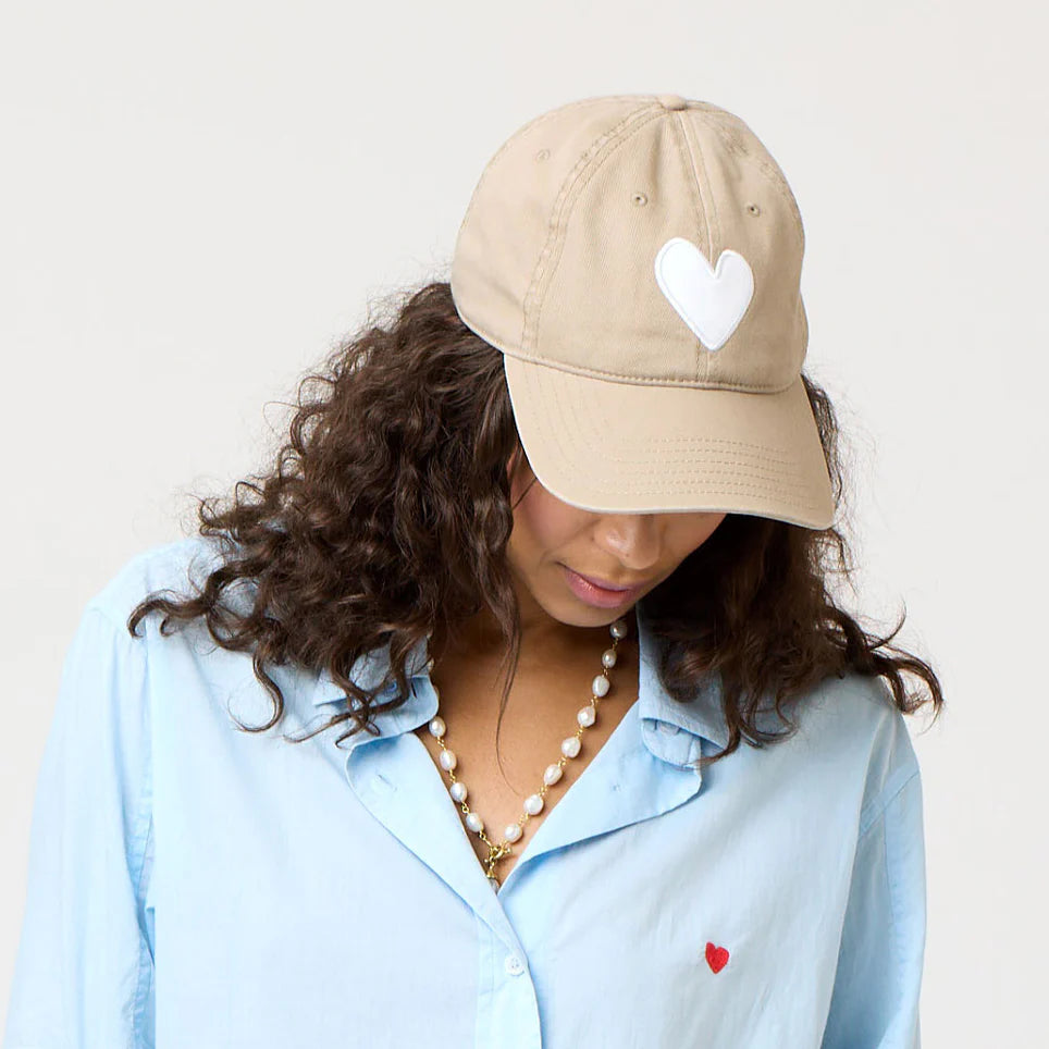 Wearing a Kerri Rosenthal KR Imperfect Heart Hat with a white heart emblem, a person looks down, revealing curly hair under the beige cap. They sport a light blue shirt adorned with a red heart and wear a pearl necklace against a plain backdrop—an exclusive collector's style.