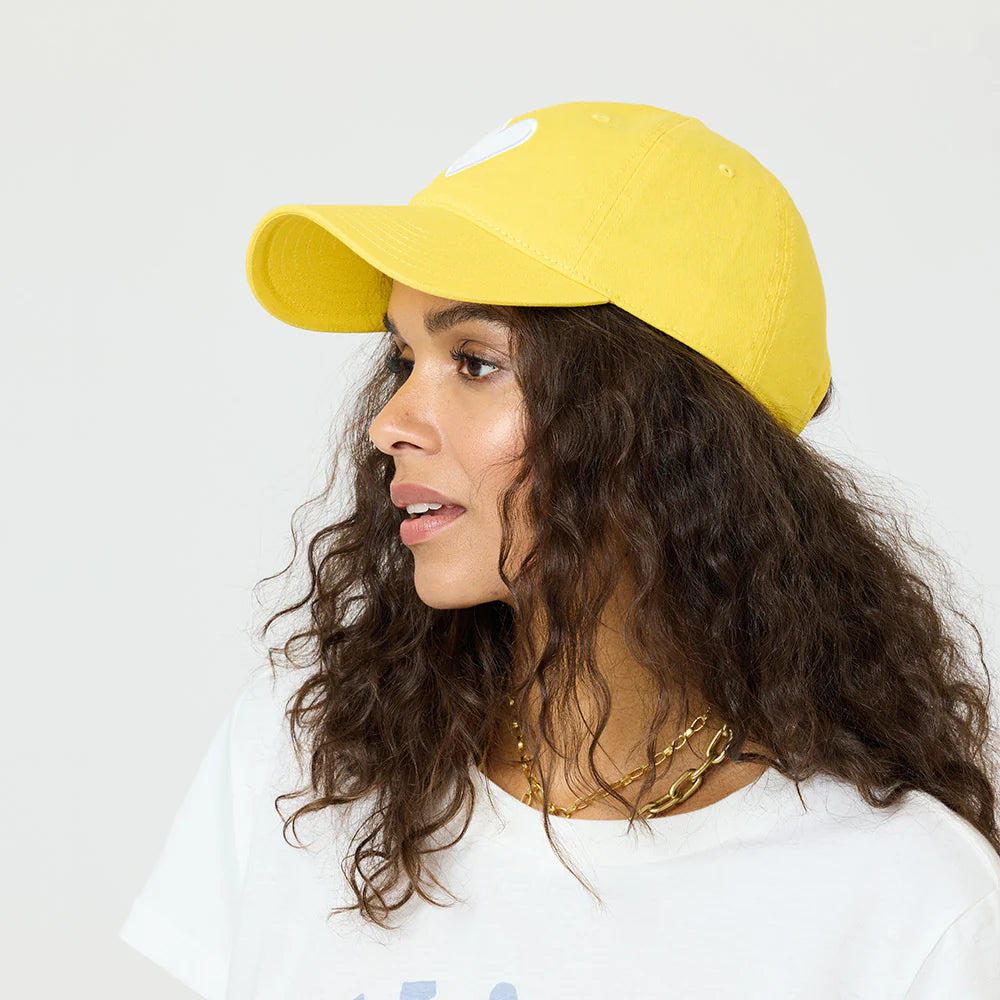 A woman with long curly hair wears a yellow KR Imperfect Heart Hat by Kerri Rosenthal, featuring an embroidered tonal heart patch, with a white t-shirt and layered gold necklaces, standing against a plain light background.