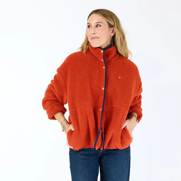 A woman stands against a white background, wearing the Kerri Rosenthal Coze Magoze cotton fleece jacket with blue trim and snap buttons, paired with dark blue jeans. She looks to the side, hands in her jacket pockets.