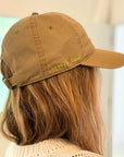 A person with light brown hair wears the Kerri Rosenthal Baseball Hat Heart Patch in tan, featuring "lovers lane" and a yellow heart embroidered on the side. The trendy hat is worn indoors, viewed from behind.
