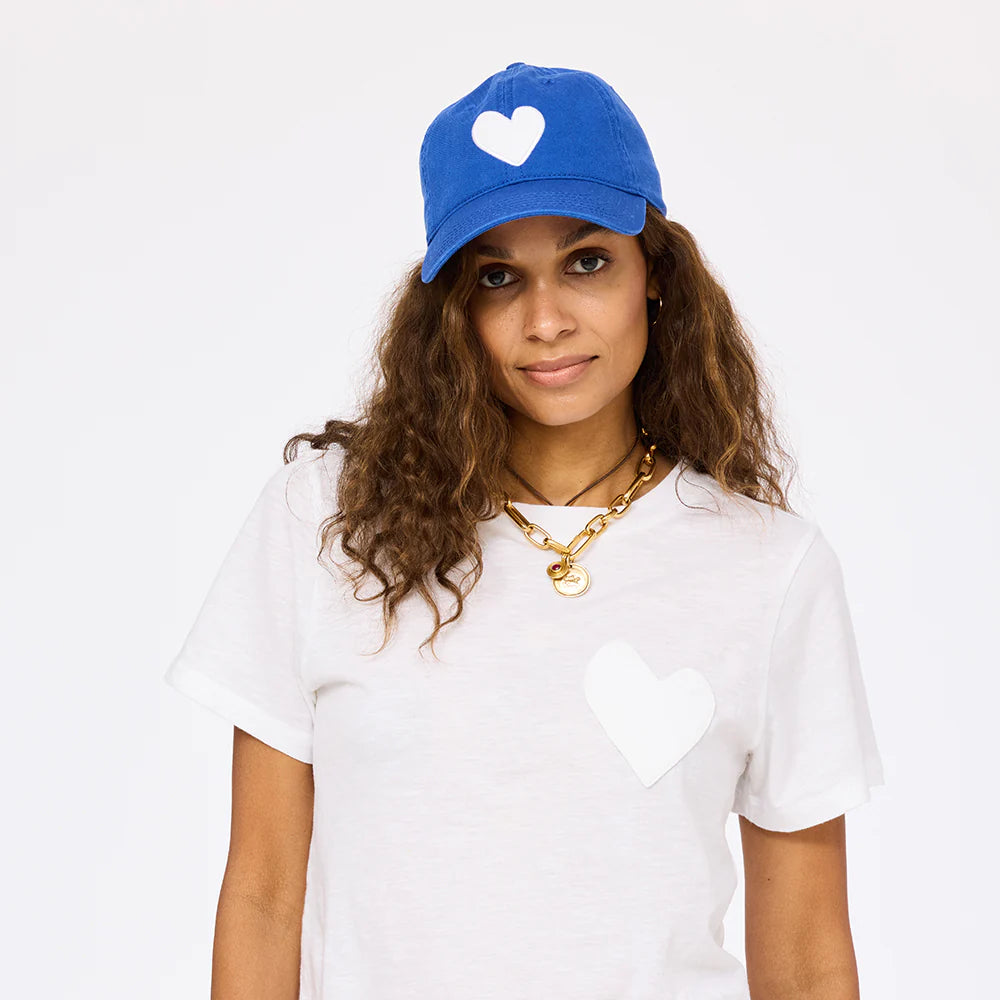 A woman with long, curly hair wears the Kerri Rosenthal KR Imperfect Heart Hat, a blue baseball cap with an embroidered heart patch, and pairs it with a gold chain necklace against a plain white background.