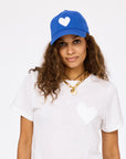 A woman with long, curly hair wears the Kerri Rosenthal KR Imperfect Heart Hat, a blue baseball cap with an embroidered heart patch, and pairs it with a gold chain necklace against a plain white background.