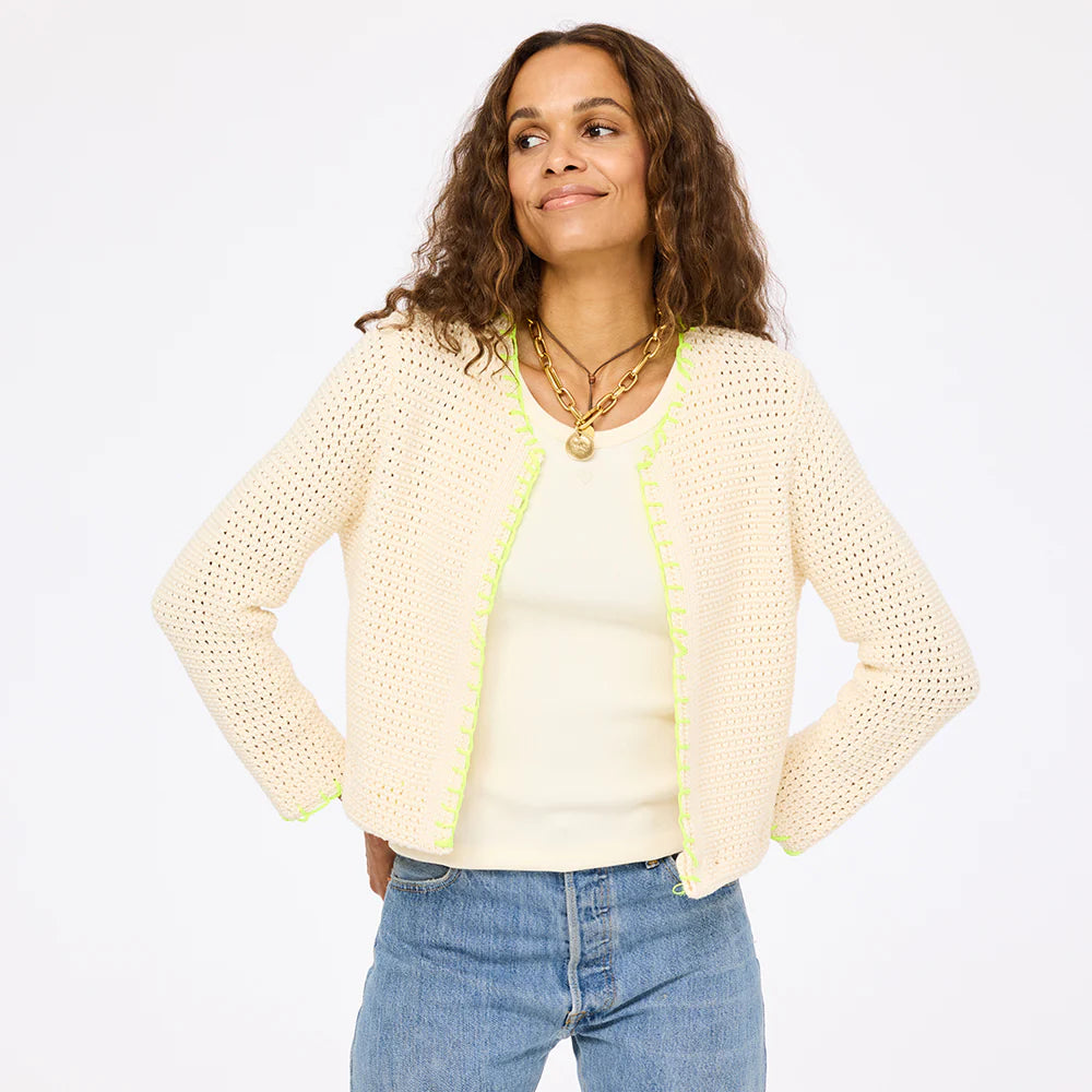 A woman with long curly hair wears the Frankie Beau-Lero open front cotton crochet cardigan by Kerri Rosenthal, featuring light yellow trim and subtle heart detail, over a cream top and blue jeans, posing against a plain white background.