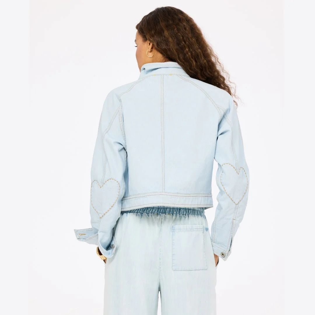 A person with long, curly hair models the Kerri Rosenthal Denim Jacket—light blue, double breasted, with heart elbow patches—and matching denim pants. They are shown from behind against a plain background.