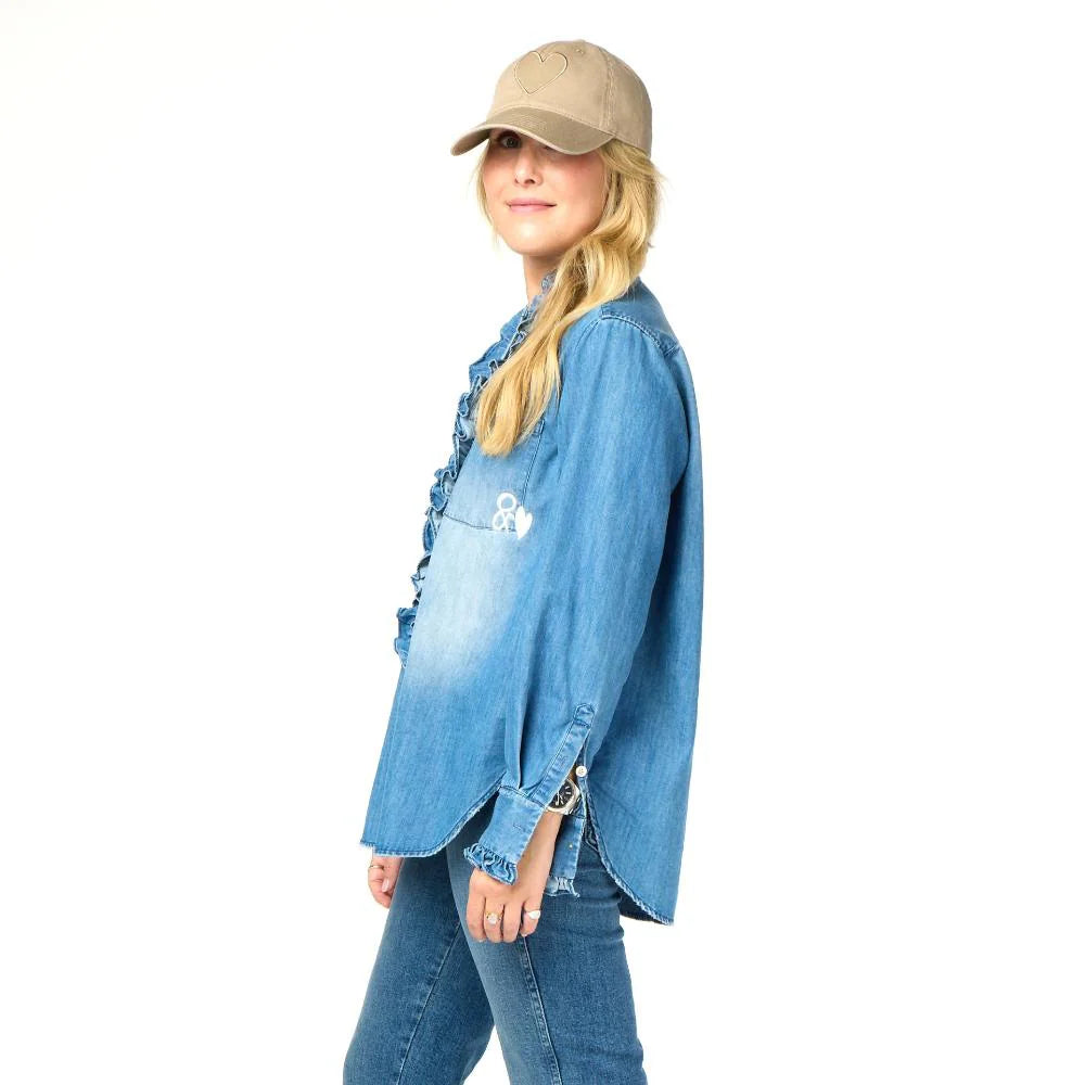 A person in a blue denim shirt and jeans stands sideways, smiling. Their long hair flows from beneath a beige hat, effortlessly stylish against a plain white background, reminiscent of the KR Imperfect Heart Hat by Kerri Rosenthal.