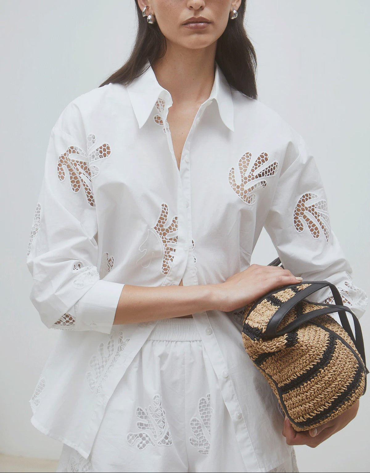 A woman wears the SUNCOO Loula Blouse, a white organic cotton button-up with cut-out details, paired with a matching skirt and holding a woven straw bag with black handles against a plain light background.