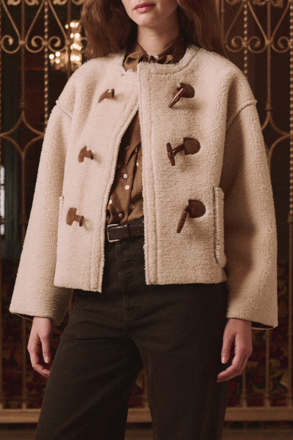 The Great Inc. Woodside Toggle Coat in brown, styled over dark pants and shirt, worn indoors for a modern look.