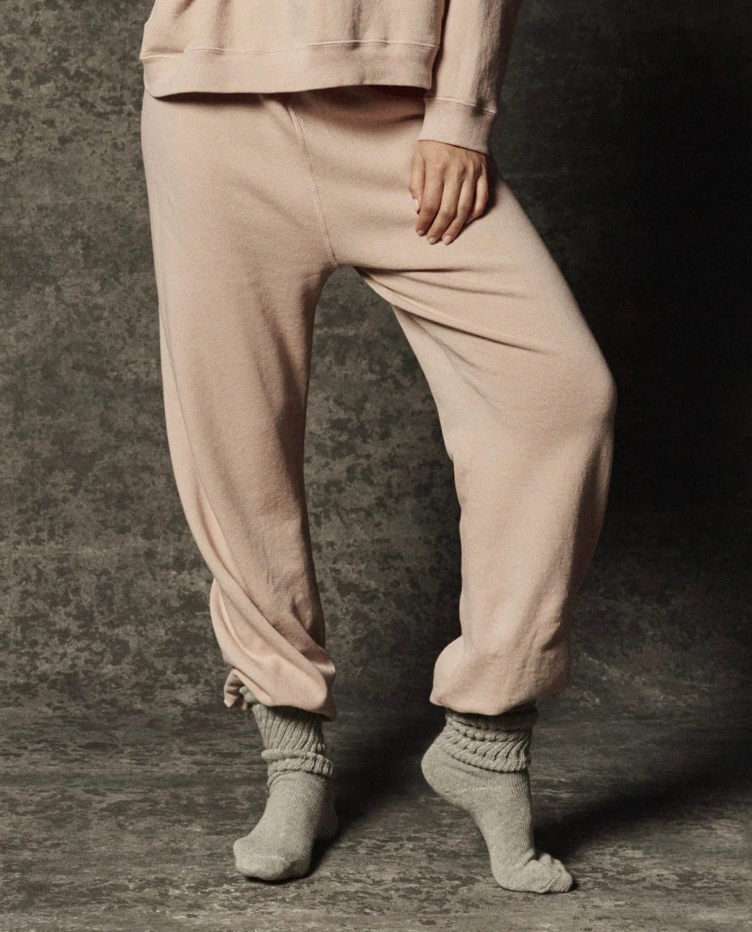 The Great Inc. Dance Pant in dark fabric, worn with gray socks, model stands on a textured gray floor.