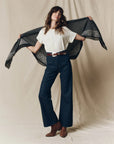 A woman stands against a beige backdrop, wearing a white tee, high-waisted dark blue wide-leg jeans, brown belt and ankle boots, holding The Big Bandana by The Great Inc. open behind her shoulders.