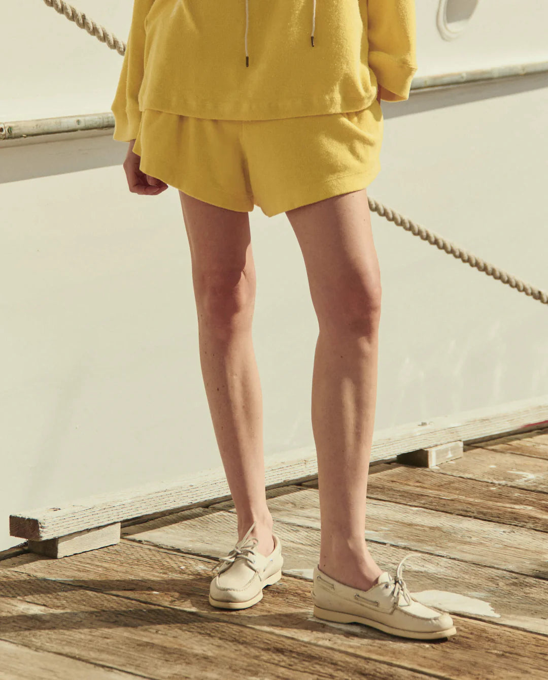 Standing on a wooden dock, a person wears The Great Inc.'s yellow Micro Terry Swing Short made from cotton micro terry and cream boat shoes. Their upper body is mostly out of frame, with a boat and rope visible in the background.