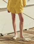 Standing on a wooden dock, a person wears The Great Inc.'s yellow Micro Terry Swing Short made from cotton micro terry and cream boat shoes. Their upper body is mostly out of frame, with a boat and rope visible in the background.
